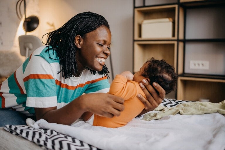 A woman is smiling while gently holding and looking at a baby who is lying on a bed. The woman, donning a colorful striped shirt, glows with the joy of motherhood as she debunks postpartum myths. The baby, dressed in an orange outfit, rests peacefully. Shelves and a lamp can be seen in the background.