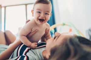 A smiling baby is being held above by an adult, who is lying down and looking up at the baby. They both appear to be playing and enjoying each other's company. Perhaps they're already thinking of H names for boys, illuminated by the bright natural light coming through a window in the blurred interior background.