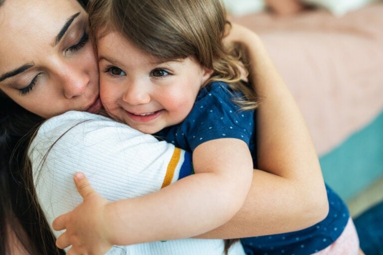 A woman and a young child share a warm hug, embodying the lessons we can learn from our children. The woman, eyes closed, holds the child close. The child, wearing a blue shirt with white polka dots, smiles brightly while embracing the woman. Both appear to be indoors in a cozy environment.
