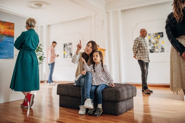 A woman and a child sit on a padded bench in an art gallery, with the woman pointing and the child looking in the same direction. Other visitors are engaging in an adventure hunt among the artworks displayed on the walls. The room has a bright atmosphere with wooden flooring.