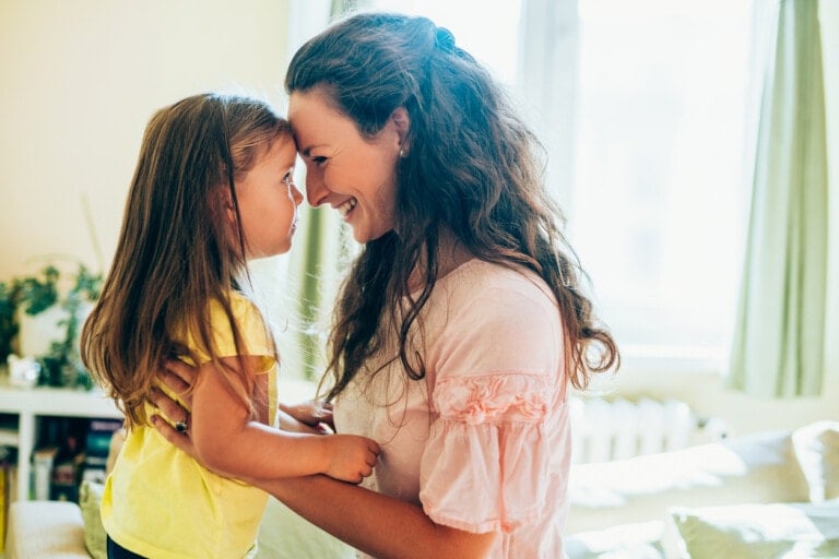 A woman with long, wavy hair is holding a young girl with long, straight hair. Both are smiling and touching foreheads. The woman is wearing a light pink blouse, and the girl is dressed in a yellow shirt, ready for preschool. They are indoors with a window in the background.