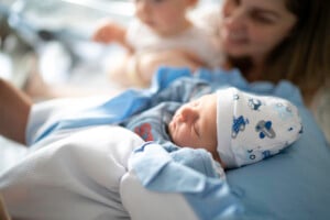 A newborn baby lies sleeping, wrapped in a blue blanket and wearing a hat with cartoon designs. The baby, perhaps destined for one of those timeless Biblical boy names, is held by an unseen person, with a blurred figure of a woman smiling and looking at the baby in the background.