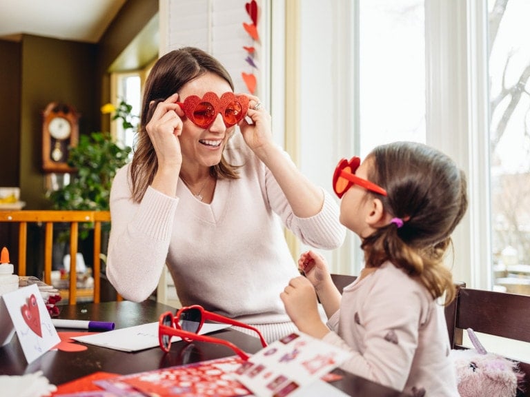 A woman and a child are seated at a table covered with crafts, both donning Valentine's Day outfits complete with heart-shaped glasses. They smile at each other warmly in the room filled with natural light from large windows, surrounded by an array of colorful craft materials.