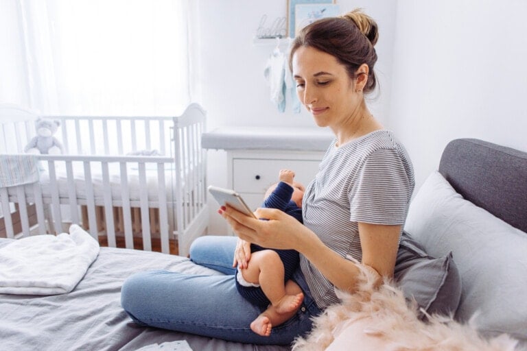 A woman is sitting on a bed, holding a baby in her lap while looking at her phone, possibly checking tracking apps. The room is bright with natural light, and a white crib is visible in the background. The woman is casually dressed, and the scene suggests a calm and relaxed moment.