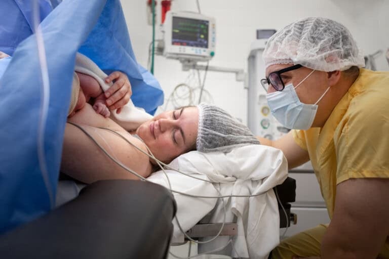 A woman lies in a hospital bed in the operating room holding her newborn baby on her chest. She and a person beside her, both wearing hairnets, are in a medical setting with monitors and equipment visible. The partner or anesthesiologist is wearing a mask and scrubs.