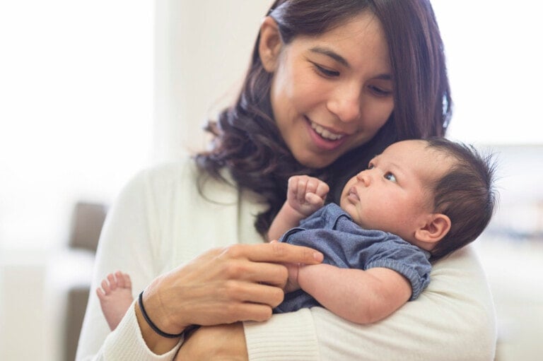A woman with long dark hair smiles while holding a baby dressed in a blue outfit, possibly pondering Mexican boy names. The baby looks up at the woman with a curious expression. The background is softly lit, suggesting an indoor setting.