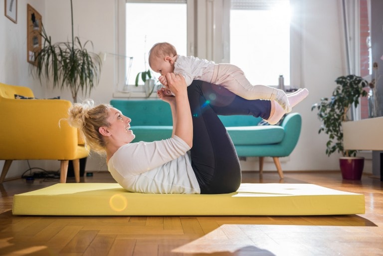 A woman, working on her postpartum weight loss, lies on an exercise mat holding a baby in the air with her hands and bent legs. The room features a yellow armchair, a blue couch, and several plants. Sunlight streams through the windows, illuminating the scene.