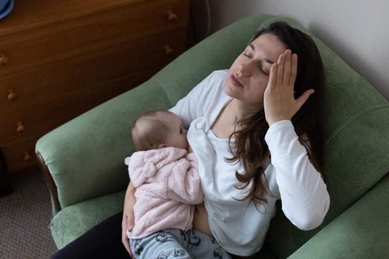 A woman sits in a green armchair, holding a baby wrapped in a pink blanket to her chest with one arm. Her eyes are closed, and she is touching her forehead with her other hand, suggesting fatigue or stress common in postpartum depression.