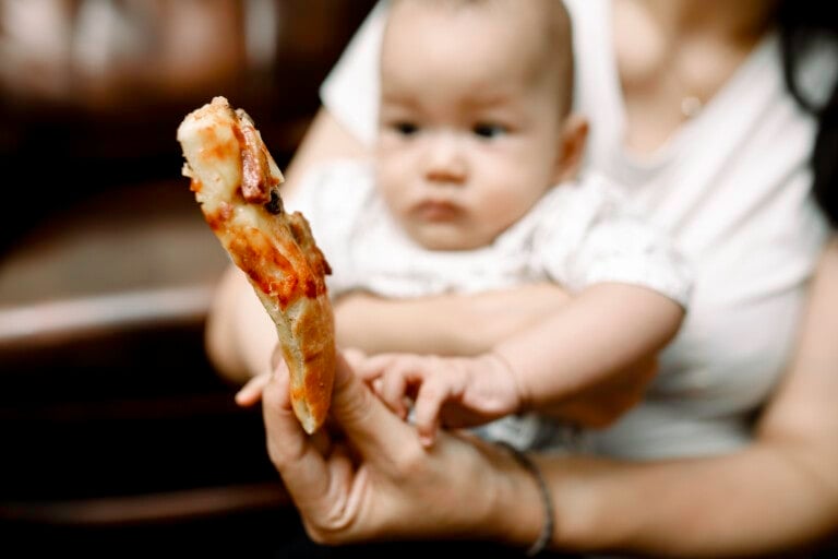 A baby, held by a person in a white shirt, looks intently at a slice of pizza with toppings, which is being held close to it by another hand. The baby appears curious and reaches out toward the pizza, showing an early interest in first foods. The background is blurred.