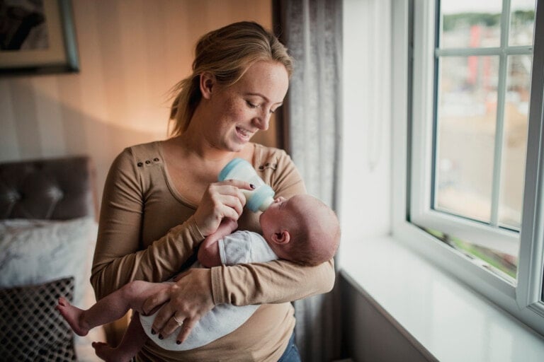 A woman is standing by a window, smiling down at a baby she cradles in her arms. She is holding a bottle to the baby’s mouth, feeding the baby with love and care. The room, filled with soft lighting and baby essentials, has a bed and cushions visible in the background.