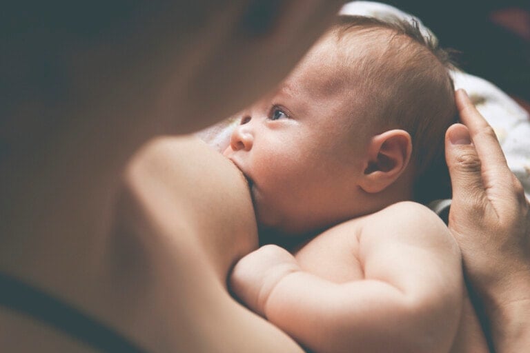 Close-up of a baby breastfeeding. The baby, who has light-colored hair, is latched onto the breast and looking up with one hand resting on the mother's chest. The mother holds the baby gently with one hand supporting the baby's head, ensuring both are comfortable as she provides essential vitamin D while breastfeeding.