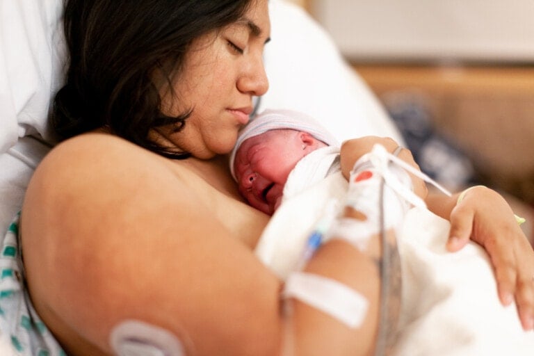 A woman lying in a hospital bed is holding a newborn baby against her chest. She has closed eyes, and the baby is wrapped in a white blanket with a cap on its head. Medical tubes and monitors are visible attached to them, highlighting the crucial care not to do after birth.
