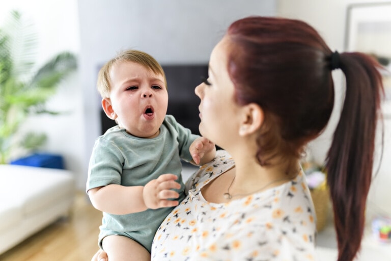 A woman holds a crying baby indoors. The baby, dressed in a green shirt, is holding a hand near the woman's shoulder and appears to be coughing. The woman, with her hair tied back in a ponytail, looks at the baby with concern, pondering if infant cough medicine might help. The background includes a plant and furniture.