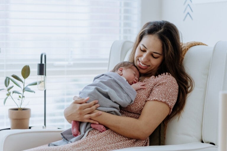 After you have a baby, a woman sits on a white chair in a brightly lit room, holding her sleeping infant on her chest. She is smiling and looking down at the baby. A small potted plant and a lamp are visible in the background.