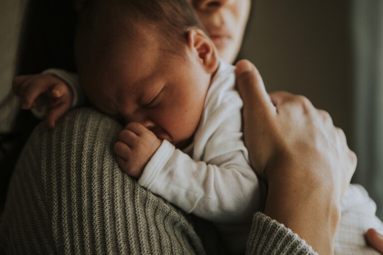 A baby in a white onesie rests its head on a person's shoulder. The person, wearing a textured knit sweater, holds the baby securely. The background is blurred, focusing attention on the baby and the comforting embrace—a tender moment of becoming a mom.