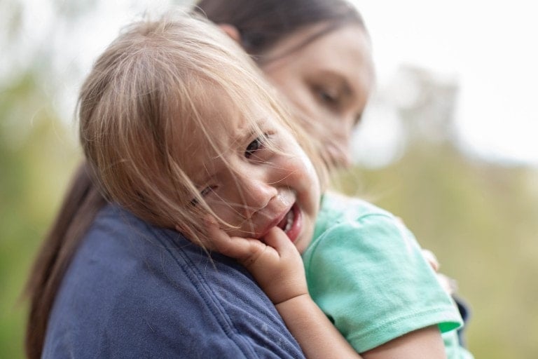 A young child, recovering from a tantrum, is being comforted by an adult who holds them in a soothing embrace. A tear glistens on the child's cheek as their face rests against the adult's shoulder and a small hand clasps their own cheek. The background is gently blurred, emphasizing their tender moment.