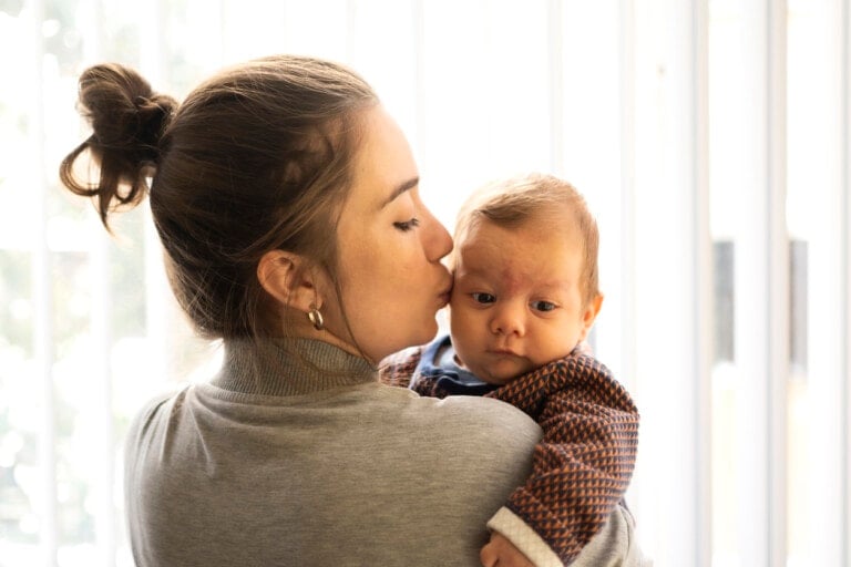 A woman with her hair in a bun kisses a baby on the cheek. The baby, wearing a patterned sweater, looks towards the camera. They are indoors with a bright background, sharing tender mom lessons.