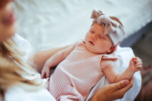 A baby wearing a pink headband with a large bow is sleeping in the arms of an adult. The baby girl, dressed in a pink, ribbed outfit, appears to be peacefully resting. The background shows part of a bed with a light-colored duvet. It's moments like these that inspire heartwarming baby girl quotes.