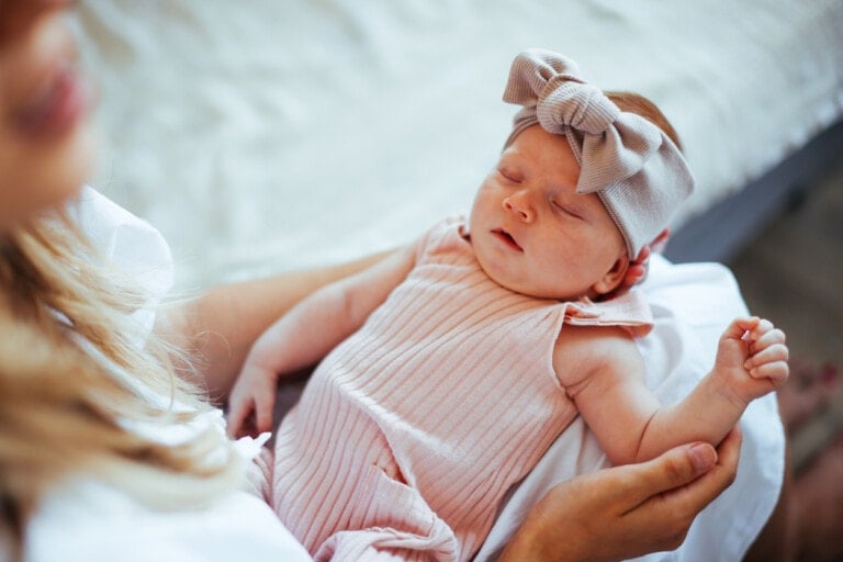A baby wearing a pink headband with a large bow is sleeping in the arms of an adult. The baby girl, dressed in a pink, ribbed outfit, appears to be peacefully resting. The background shows part of a bed with a light-colored duvet. It's moments like these that inspire heartwarming baby girl quotes.