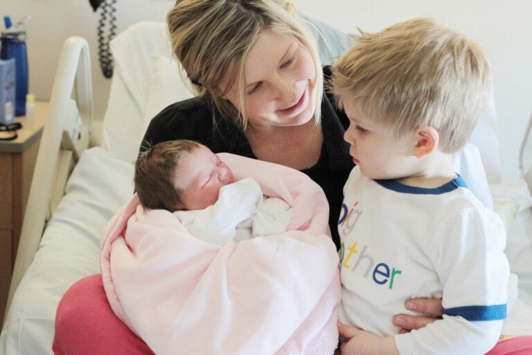 A woman in a hospital bed holds a newborn wrapped in a pink blanket. She smiles at a young boy standing next to her wearing a shirt that reads "Big brother". The boy looks at the baby with curiosity, as the mother embraces being postpartum again.