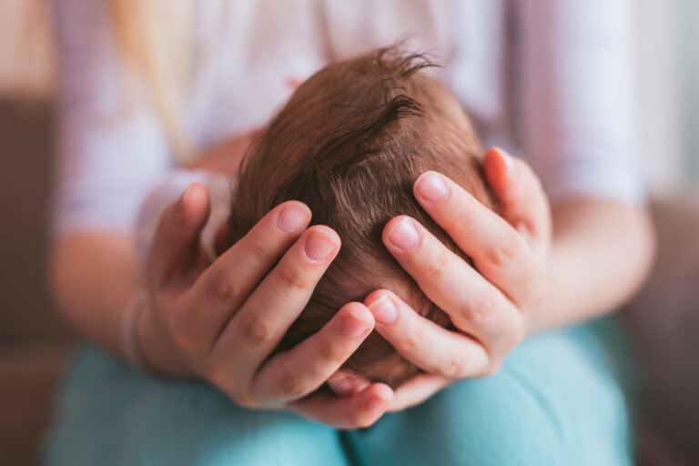Close-up of a person's hands gently cradling a baby's head during the baby's first night home. The baby's dark hair is visible, and the background is blurred. The person is wearing a white long-sleeve top, and the baby is wrapped in a blue blanket.