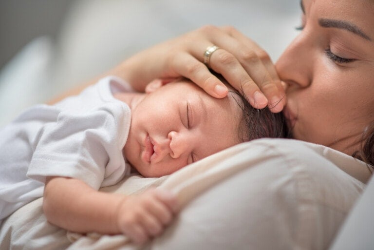 A person with long hair is lying down, holding and kissing the forehead of a sleeping baby dressed in a white outfit. The baby, enveloped in the warmth of the fourth trimester, rests on the person's chest, with one hand gently touching the fabric.