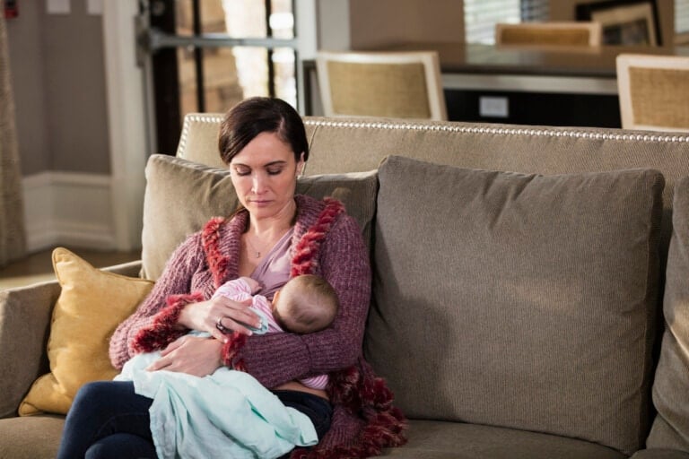 A woman is sitting on a couch, cradling a baby wrapped in a blanket in her arms. She looks serene as she embraces the "breast is best" philosophy. The cozy living room has beige and yellow pillows on the couch, with a dining area and chairs visible in the background.