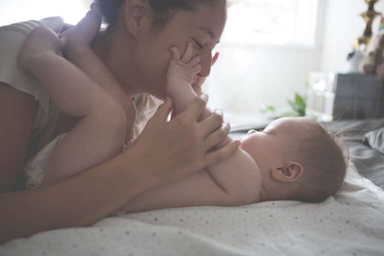 A person lies on a bed with a baby, touching their face lovingly. The baby is lying on a soft surface, reaching up and touching the person's face. Softly lit with natural light streaming through a window in the background, this serene scene captures the essence of tender moments and offers advice for new parents: cherish every second.