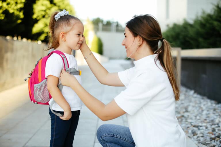 A mother kneels and adjusts the shoulder strap of a pink backpack worn by her daughter, a young girl, with a ponytail, ready for her first day of kindergarten. They are outdoors on a paved pathway with greenery in the background. Both are smiling.