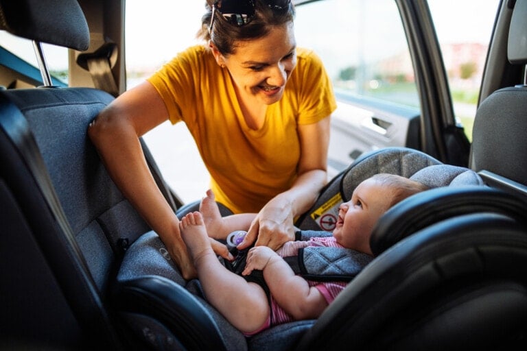 A woman in a yellow shirt is smiling as she ensures baby safety by securing a baby in a car seat inside a car. The baby, strapped in the seat, is also smiling and looking at the woman. The car door is open, and it appears to be a bright day outside.