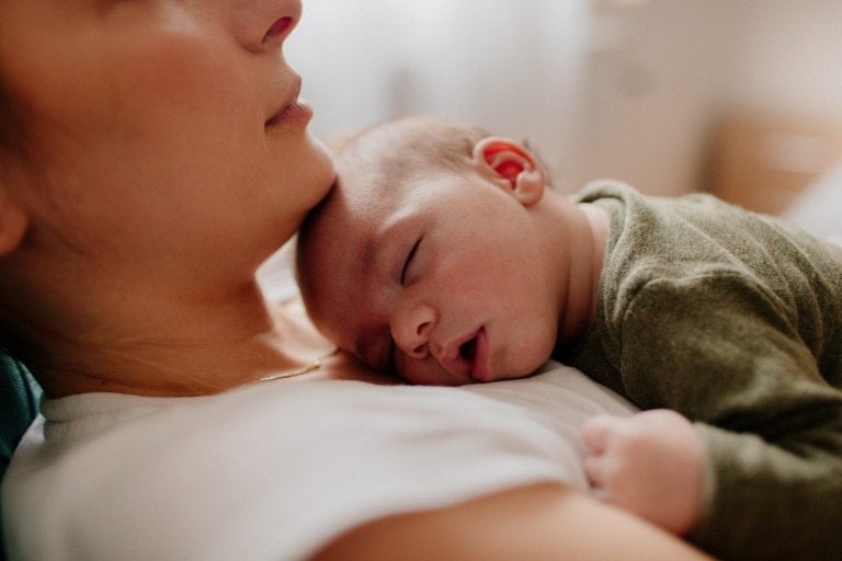 An infant is sleeping peacefully with its head resting on an adult's shoulder. The adult is wearing a white shirt, and the baby, possibly adjusting to a newborn sleep schedule, is wearing a green outfit. The background is softly blurred, suggesting an indoor setting.