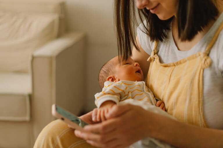 A woman dressed in a yellow jumper and white shirt is holding a newborn baby in her arms. The baby, wearing white clothes with yellow stripes, rests peacefully as the woman looks at her phone, scrolling through it. In the background, a beige armchair adds to the cozy scene.