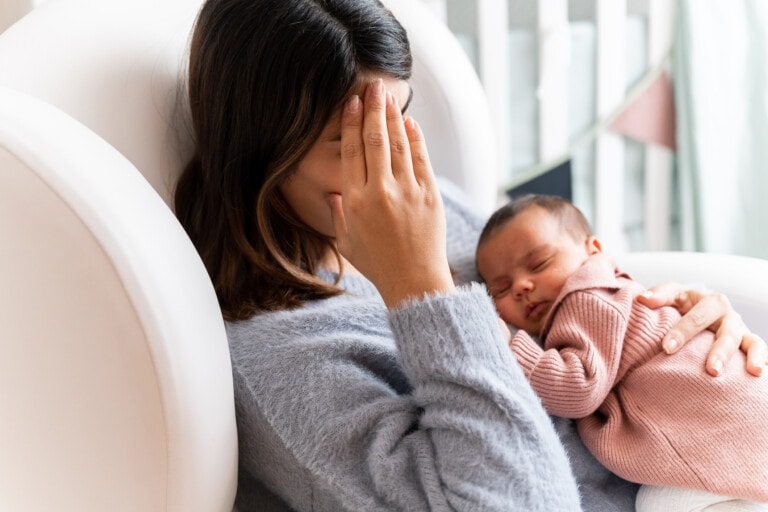 A woman sits in a white chair with one hand on her forehead, appearing tired. She is holding a sleeping baby on her chest, who is wrapped in a pink sweater. The background shows part of a crib and some hanging decorations, capturing the tender moments filled with postpartum emotions.