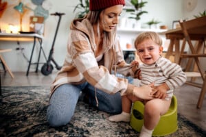 A woman wearing a red beanie and plaid jacket helps a crying toddler sitting on a green potty. The setting is a cozy, cluttered room with a scooter in the background, along with furniture and various items as the child tries to poop on the potty.