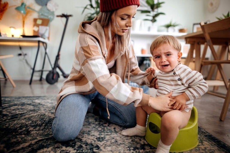 A woman wearing a red beanie and plaid jacket helps a crying toddler sitting on a green potty. The setting is a cozy, cluttered room with a scooter in the background, along with furniture and various items as the child tries to poop on the potty.