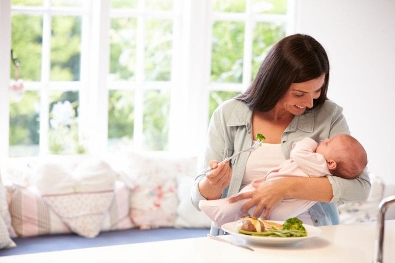 A woman is sitting at a table, practicing mindful eating with a baby in one arm and a fork in the other. On the table is a plate of vegetables. She gazes at the baby and smiles, while large windows in the background reveal an outdoor view.