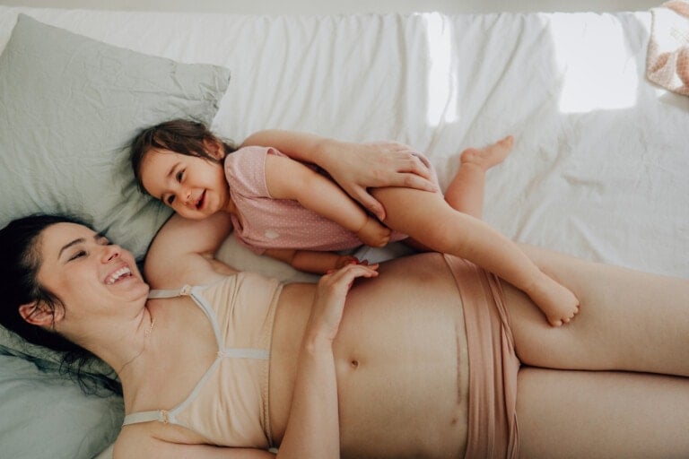 A pregnant woman, wearing beige underwear and a light bra, lies on a bed with a green pillow. A young child in a pink outfit is playfully lying on her, both smiling and looking at each other affectionately; capturing a tender moment often shared by moms who have c-sections.