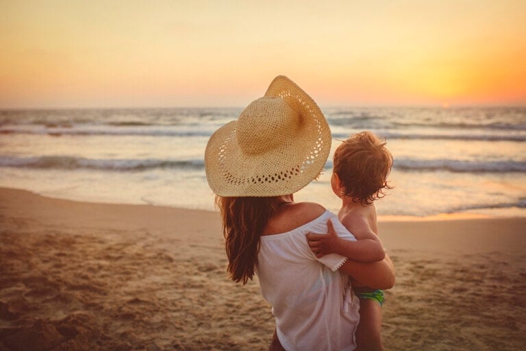 A woman wearing a straw hat is standing on the beach holding a small child while they both face the ocean during sunset. The sandy shore and gentle waves are visible, with the sun low in the sky creating an orange glow, capturing a perfect vacation moment.