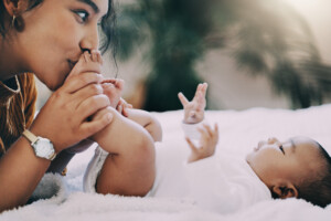 A woman is kissing the feet of a baby who is lying on a bed. The baby, with both arms raised, gazes upward. The scene exudes calm and intimacy, with a softly blurred background enhancing the tender moment of kissing baby’s tiny feet.
