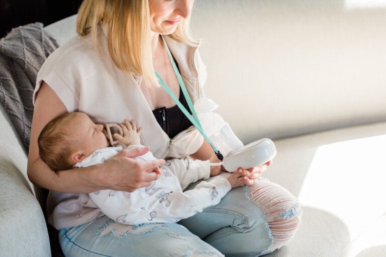 A woman is seated on a couch using an electric breast pump while holding a baby, who is dressed in a white outfit with grey patterns. The woman, wearing a light-colored shirt and light blue ripped jeans, is bathed in sunlight streaming in from the right side.