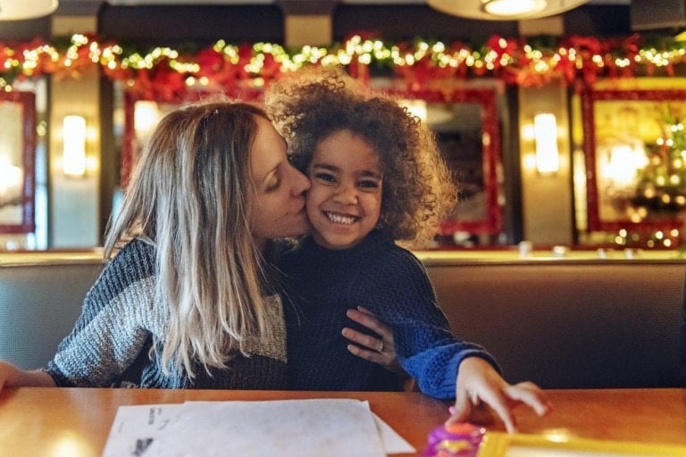 A woman kisses a smiling child on the cheek. They are seated at a table in a festive restaurant with holiday decorations, including garlands and lights, in the background. The table has papers and a candy wrapper on it, adding to the warm atmosphere often found with kids at restaurants during celebrations.