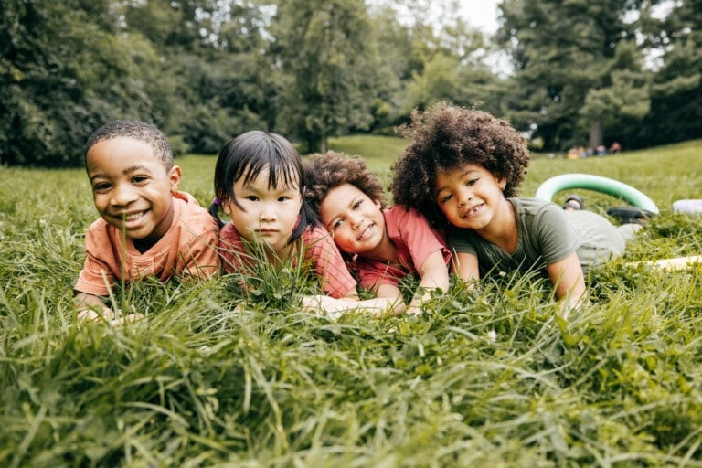 Four children lie on the grass in a park, smiling and looking at the camera. Trees and greenery are in the background, with a few people visible in the distance. The children are dressed casually and appear to be enjoying a playful spring playdate outdoors.