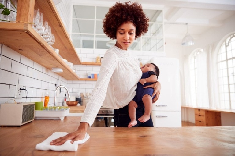 A woman with curly hair holds a baby in her left arm while she efficiently wipes a wooden kitchen counter with a cloth using her right hand. The kitchen, featuring white tile walls, wooden shelves with dishes, and a window that lets in daylight, embodies practical cleaning tips and tricks.