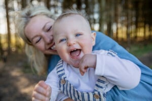 A smiling woman holding a happy baby outdoors. The baby, wearing a white shirt with striped overalls, has its mouth open in a joyful expression. The background shows blurred trees and sunlight, creating an idyllic setting for this moment. Names like those found among Celtic boy names could suit such a joyous child.