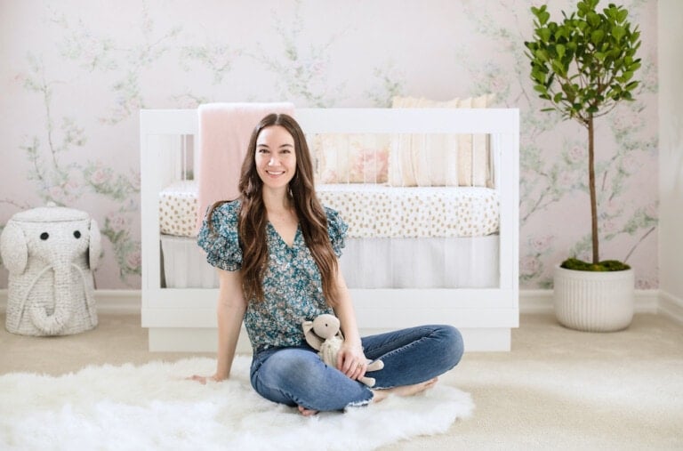 A woman with long dark hair, wearing a floral shirt and jeans, sits on a white rug holding a stuffed animal. Behind her is a beautifully designed nursery featuring a white crib with soft pastel bedding and a large green potted plant, all set against light-colored, floral wallpaper. Design your perfect nursery today!
