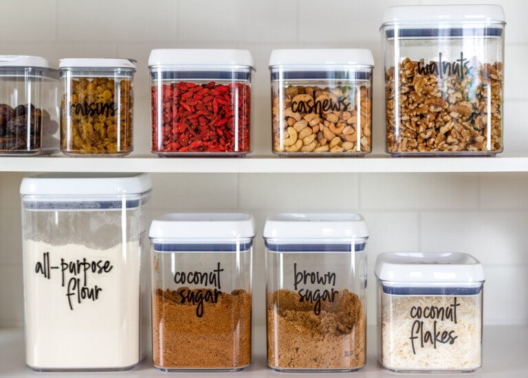 A kitchen shelf in the pantry displays clear plastic containers with white lids. Each container is labeled with its contents: raisins, dried goji berries, cashews, walnuts, all-purpose flour, coconut sugar, brown sugar, and coconut flakes—an array of superfoods lined up against a white tiled wall.