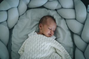 A newborn baby sleeps peacefully in a white, cushioned crib with a soft knitted blanket. The crib boasts a braided protective bumper encircling the infant.