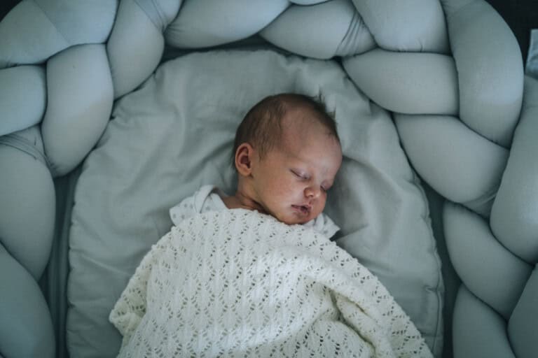 A newborn baby sleeps peacefully in a white, cushioned crib with a soft knitted blanket. The crib boasts a braided protective bumper encircling the infant.