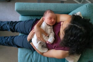 A person with curly hair is sitting on a teal couch during their postpartum recovery period while holding a sleeping newborn baby dressed in white clothing. The person is wearing a purple sleeveless top and dark pants. The scene appears calm and intimate.