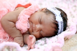 A baby wearing a frilly pink outfit and a white headband with a bow sleeps on a pink and white crocheted blanket. The baby, possibly named one of those beautiful rare girl names, lies on its side with one hand tucked under its head.